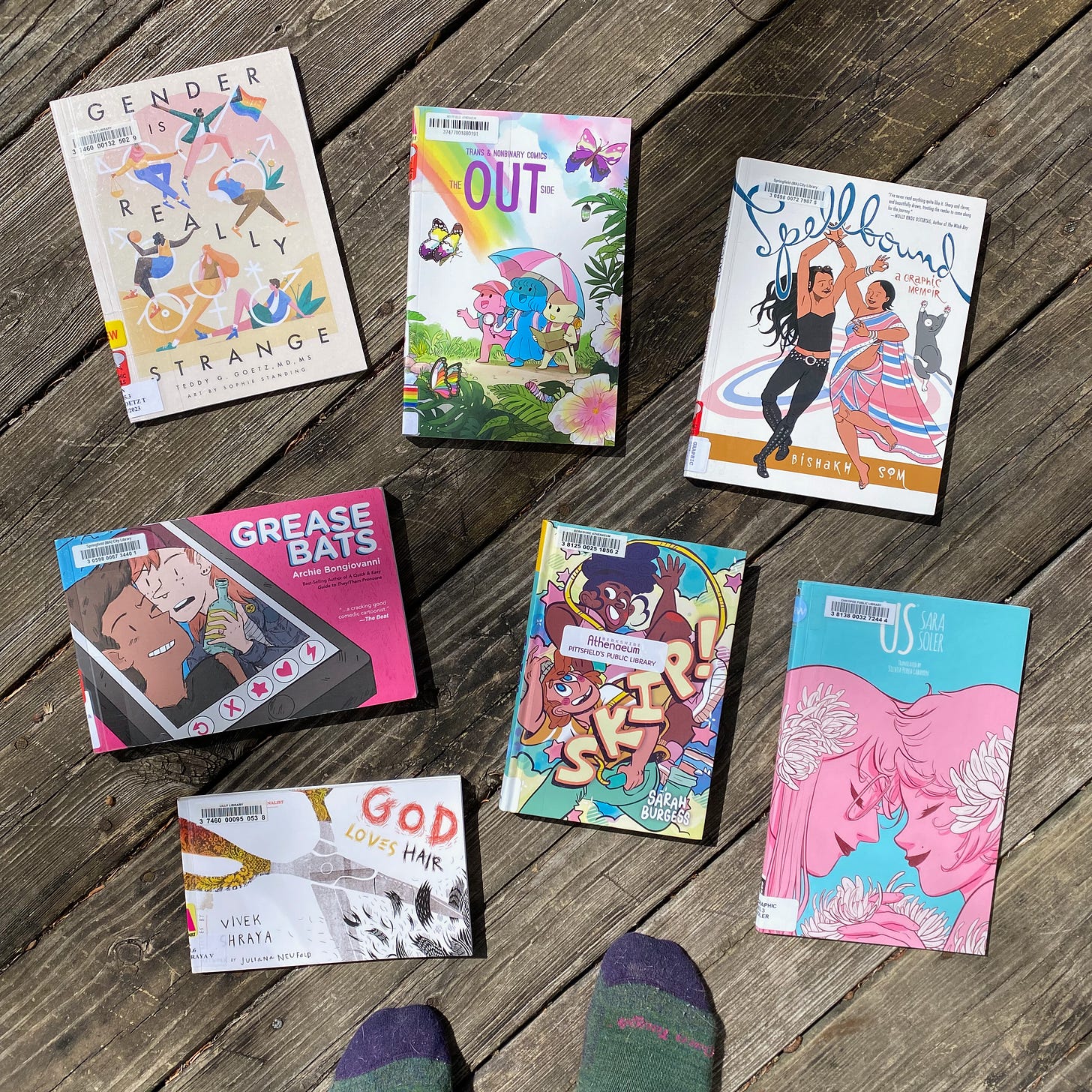 The following books spread out on a porch next to my socked feet: Gender is Really Strange, The Out Side: Trans & Nonbinary Comics, Spellbound, Us, Grease Bats, God Loves Hair, and Skip!