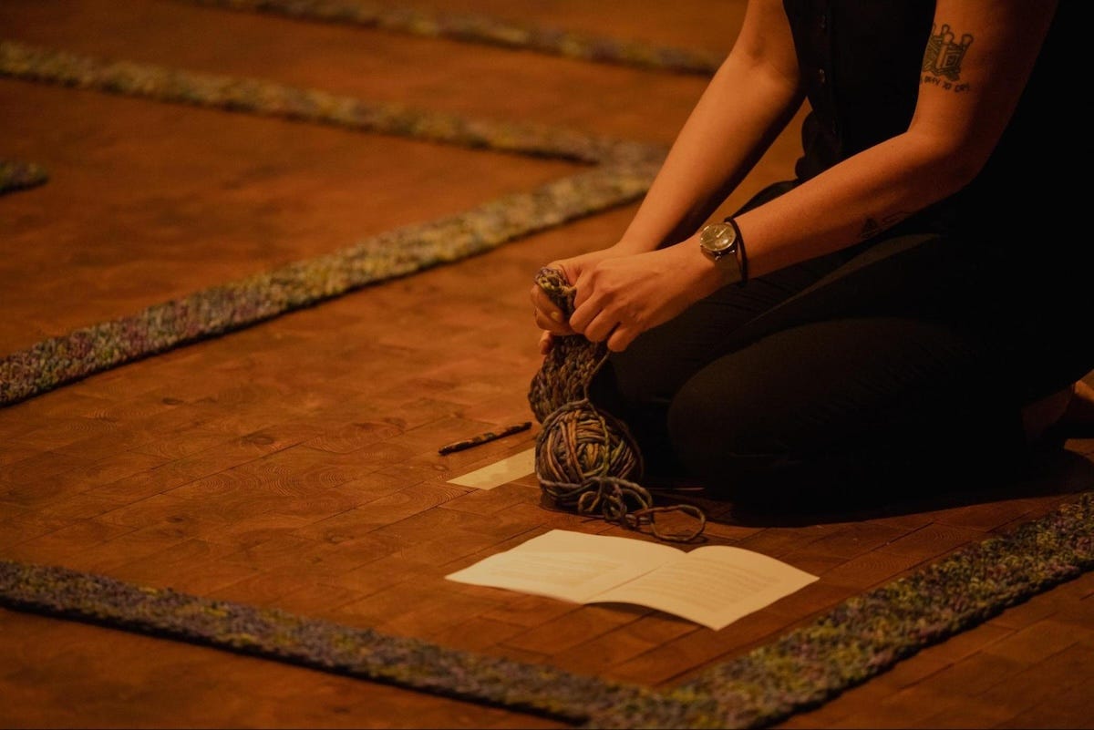 A single woman kneeling on a wooden parquet floor clutching yarn with straight knitted yarn tracks surrounding her.