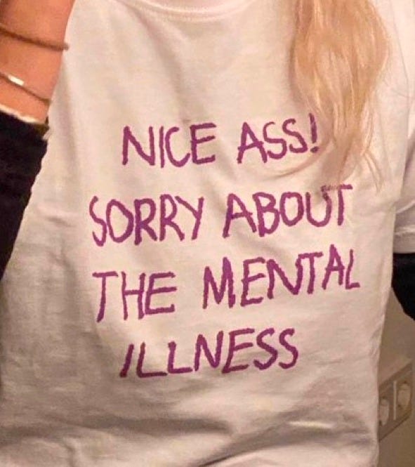 Shoulder-down photo of a white woman with blonde hair wearing a white shirt that reads in magenta font: "Nice ass! Sorry about the mental illness."