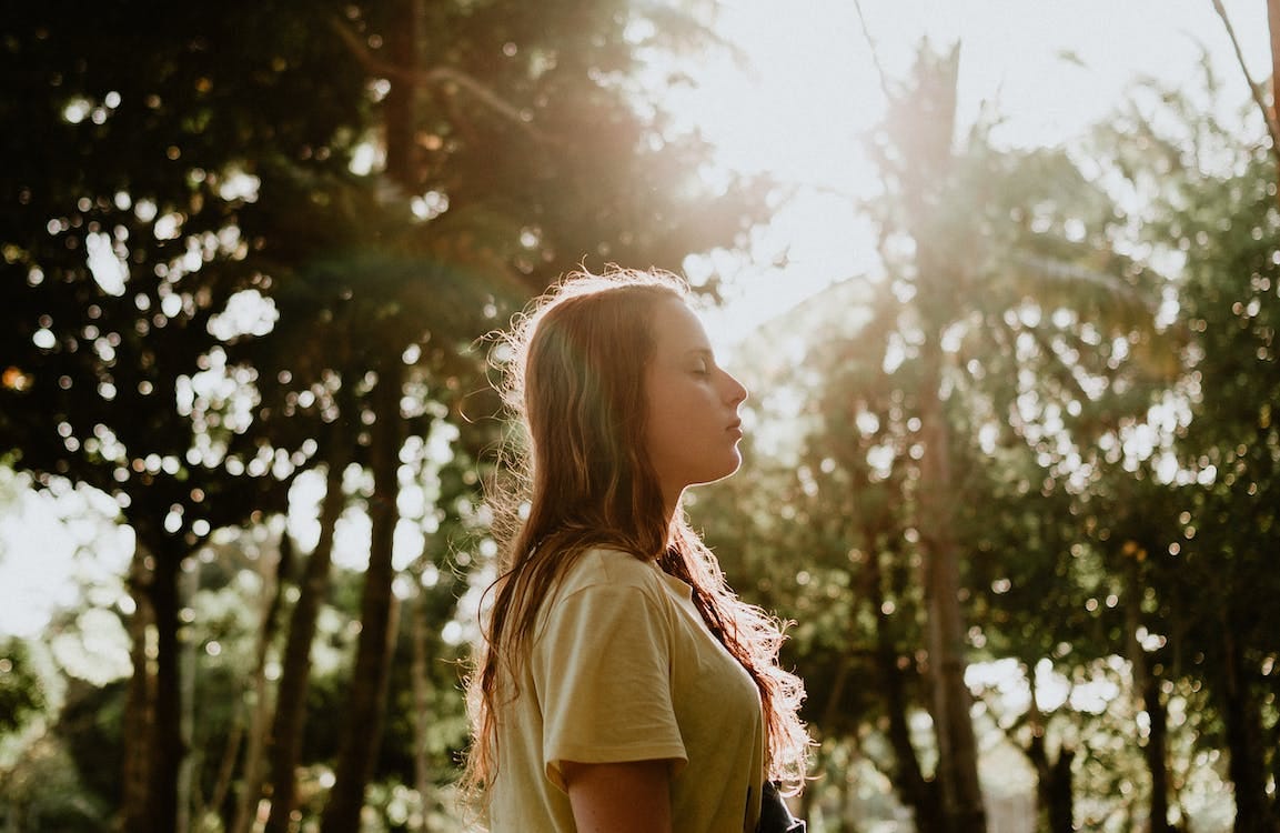 Free Side View Portrait Photo of Woman in Yellow T-shirt Standing With Her Eyes Closed With Trees in the Background Stock Photo