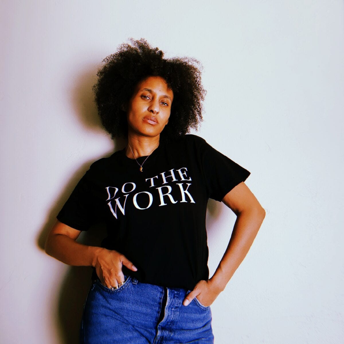 Black woman with curly black hair wears a black-and-white shirt. The white letters say DO THE WORK. A gold necklace hangs over her shirt.