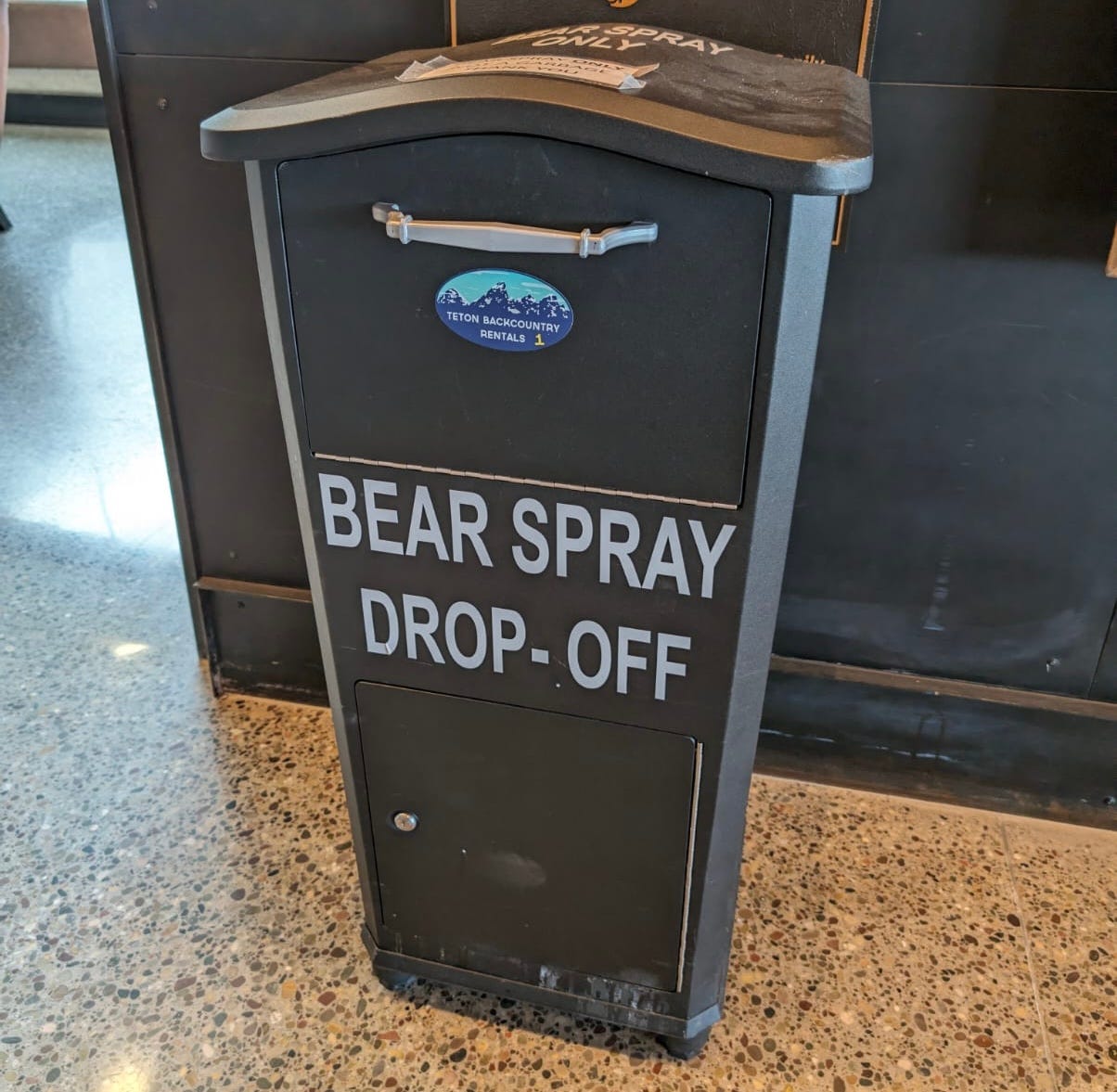 A box labeled "BEAR SPRAY DROP-OFF" with black casing, a silver handle, and a blue logo for Teton Backcountry Rentals