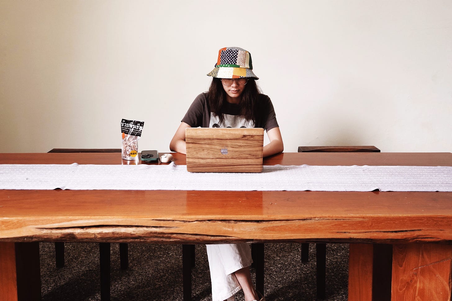 me in a patchwork hat sitting at a long wooden table with my laptop