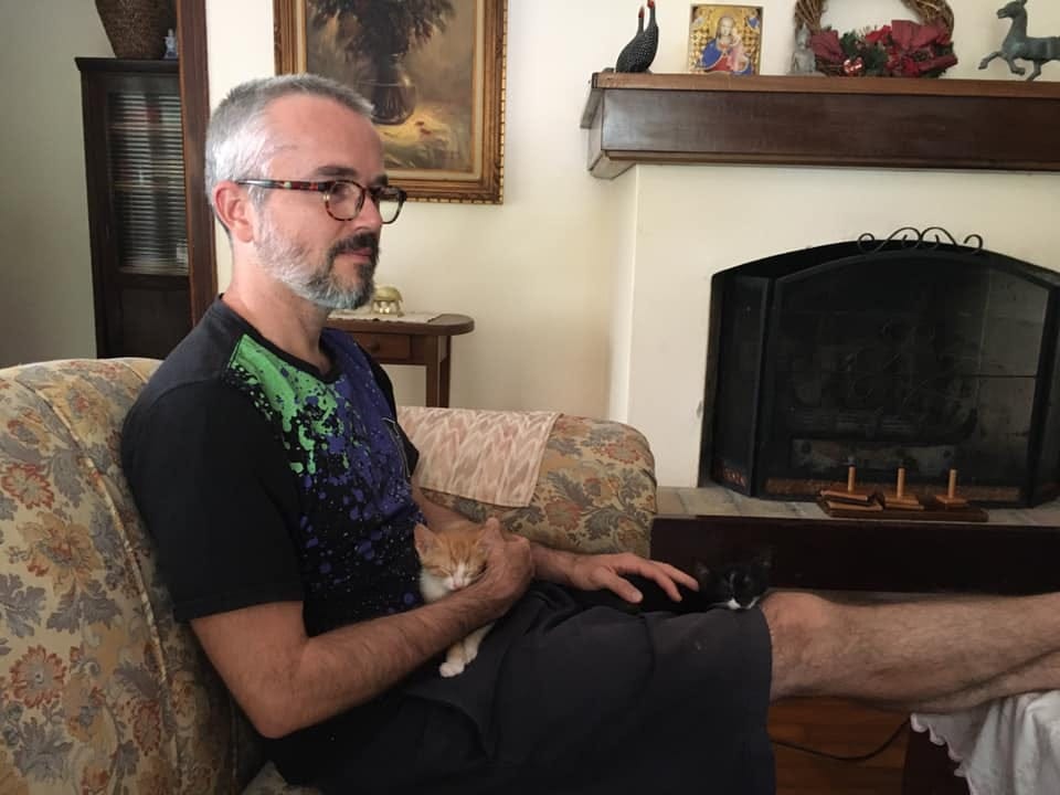 The author in his mid-forties, wearing glasses, sitting in a living room, next to a fireplace, with two kittens on his lap, a ginger one and a tuxedo one.