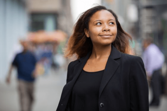 Foto de jovem negra da cintura para cima. Ela tem cabelos longos e lisos e veste blazer e blusa na cor preta. Ela olha para o lado pelo canto do olho, como quem está vendo algo além. O fundo, desfocado, mostra prédios e pessoas andando em calçada.