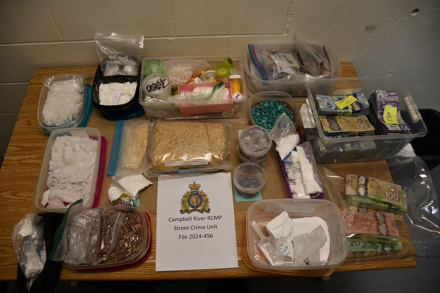 A table full of drugs in various forms, including powder, crystal, pills, in a number of different colours, as well as stacks of bills. On the table, a sheet of paper has the RCMP crest, with the words "Campbell River RCMP street crime unit file 2024-456"