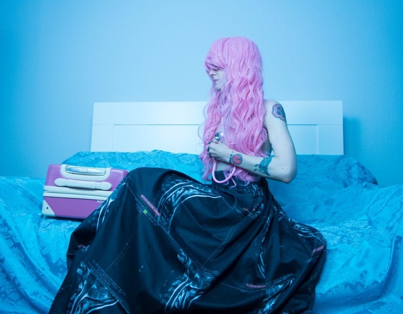 Karrie sits on a couch covered in blue damask taffeta, wearing a long, flowing skirt printed with her C-Spine MRI. She is wearing a bubble gum pink wig and holds a pink stethoscope to her heart, listening to her own heartbeat. Next to her on the couch sits a bubble gum pink suitcase, unopened.