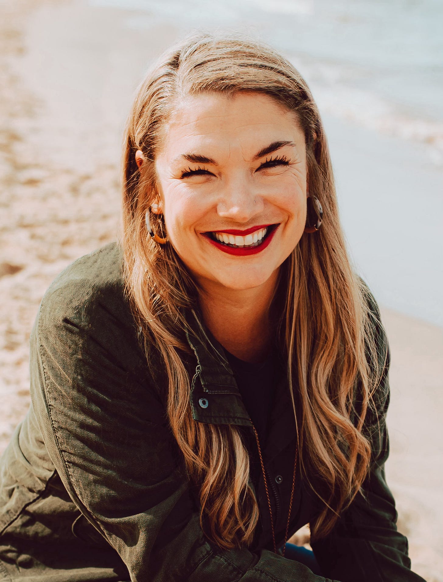 a headshot of Aundi, a white woman with a broad smile, long blond hair, with the beach and ocean behind her