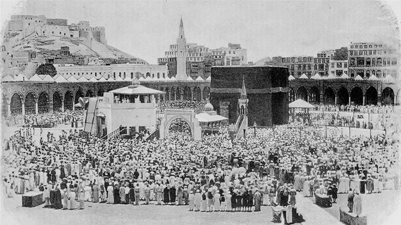 Black and white photo of the Ka'aba surrounded by tents and a few hundred pilgrims. Minarets, archways, and the hills of Mecca on which are perchedlow white buildings are visible in the background.