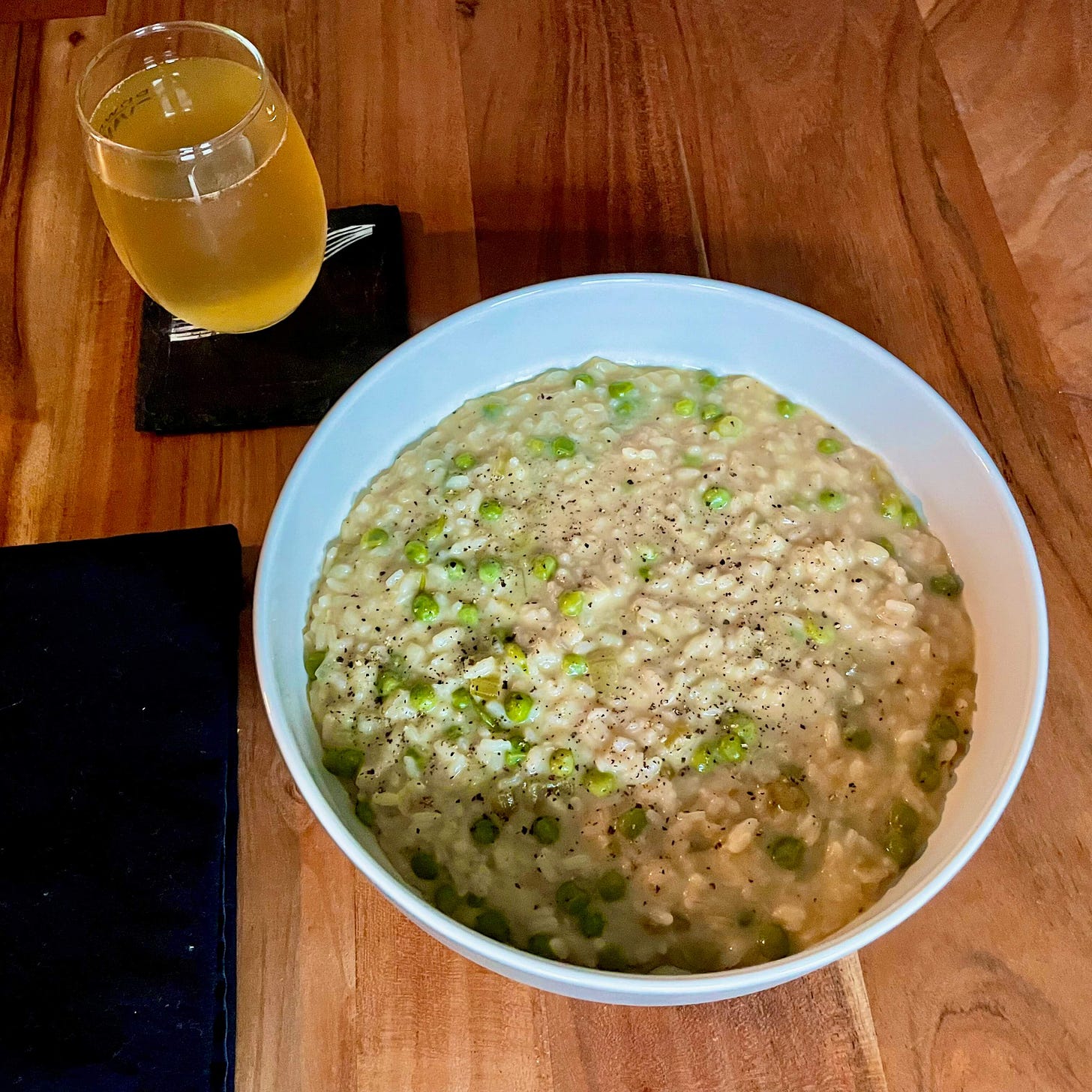 A bowl of risotto with green peas in the center of the image. To the top left is a small glass with kombucha in it.