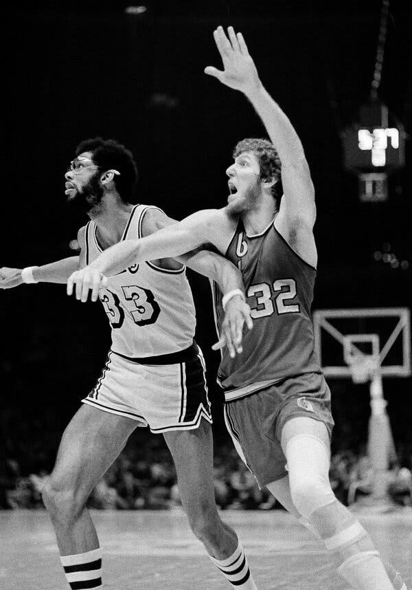 A black-and-white photo of Kareem Abdul-Jabbar and Bill Walton competing in a game, their arms reaching as they struggle for position.