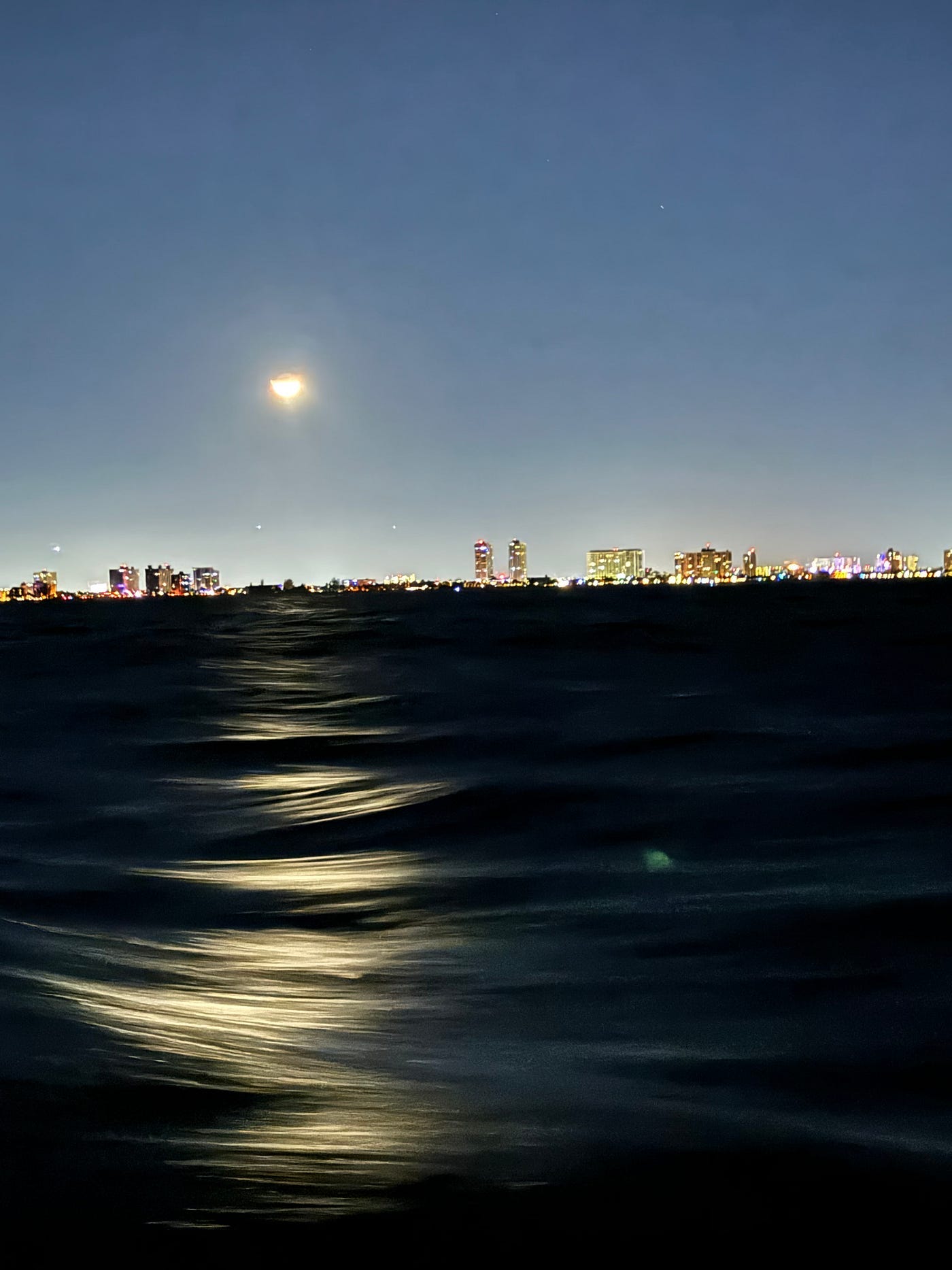 A night photo of Miami Beach skyline taken from the ocean. The reflection of of the moon is dancing on the waves.