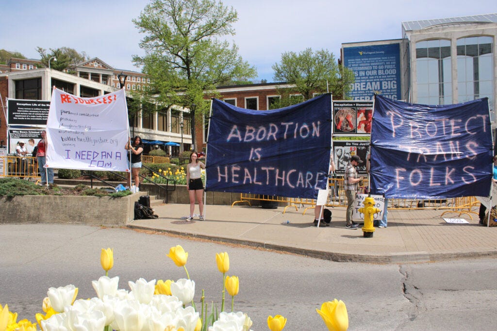 Large signs on blue tarps read "Abortion is Healthcare and "Protect Trans Folks." Two people prop up the signs on either side. The signs are held up in front of other signs, which can be seen in the gaps between the tarps, depicting what are allegedly aborted fetuses and victims of genocide. White and yellow daisies are in the extreme foreground. The entire scene takes place in front of the Mountainlair student union at West Virginia University.
