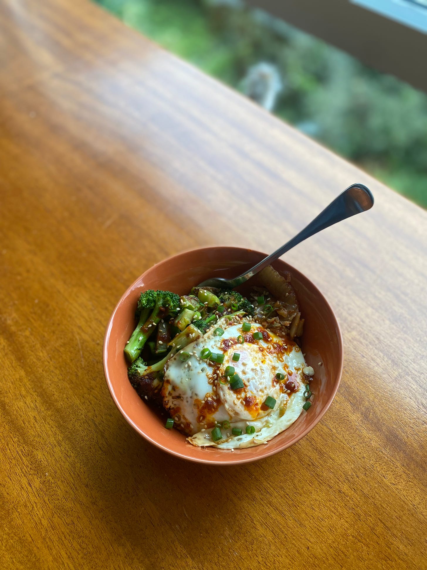 an orange bowl of the rice dish described above, a fried egg covering most of it, the broccoli and kimchi on one side. Everything is sprinkled with green onion, sesame seeds, and chili miso.