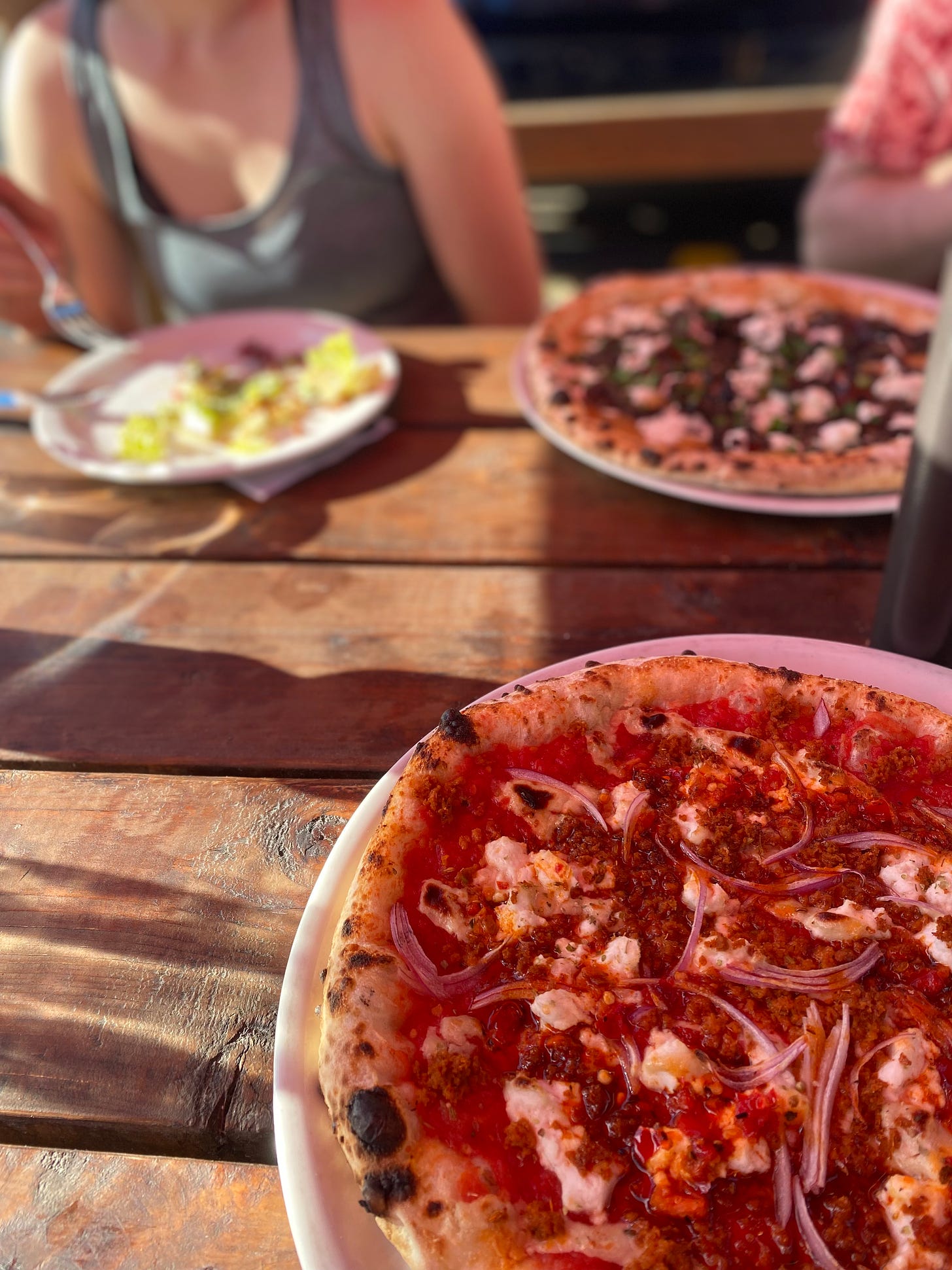 A wood patio table partially in the sun, with two pizzas on large plates across from each other. In the foreground is the bee sting, with chilies, red onion, crumbled 'meat' and cheese over red sauce; in the background, mushrooms and dollops of creamy cheese dusted with parsley. Natalie sits to the left, her plate still with a little caesar salad on it.