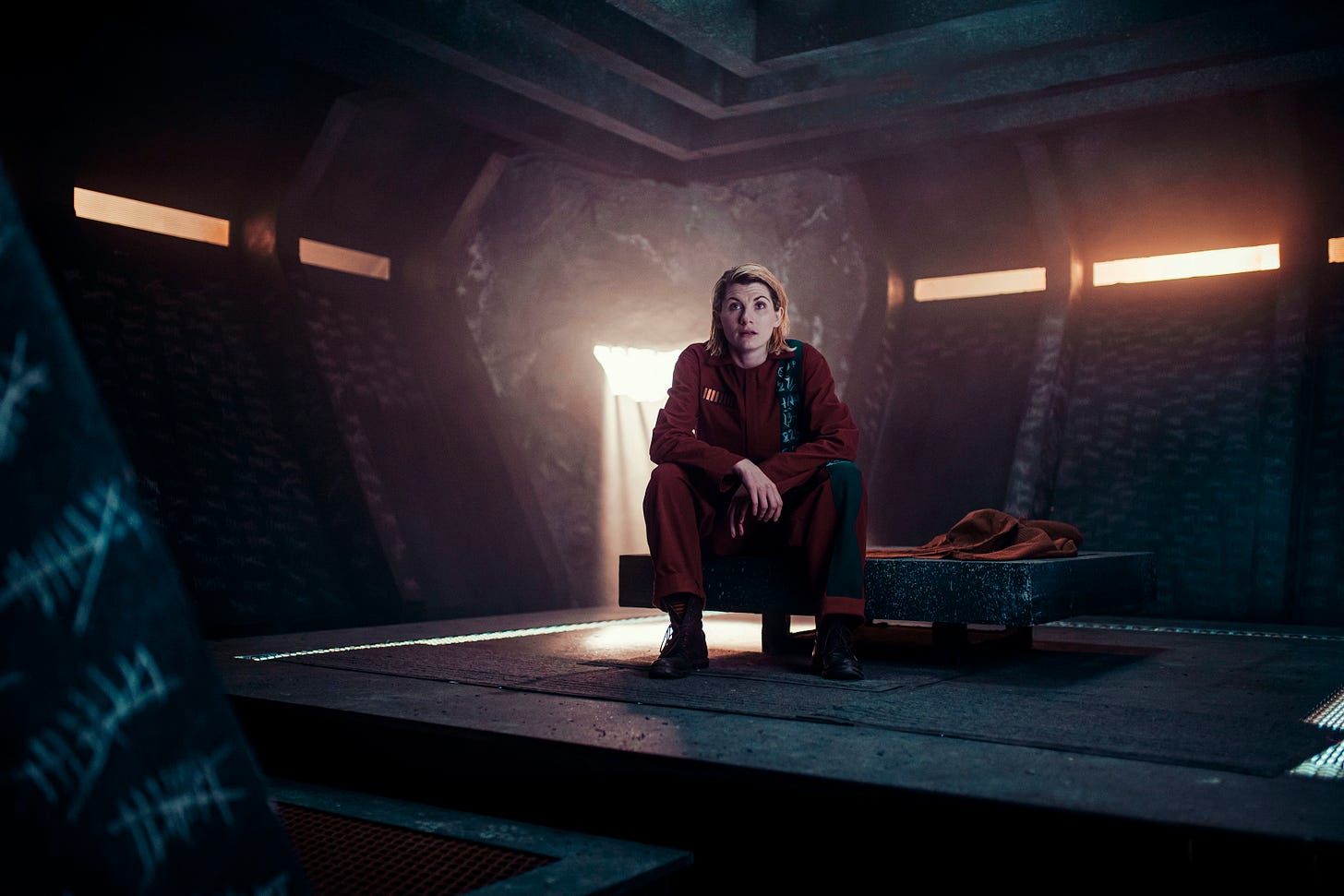 This image shows the Thirteenth Doctor (played by Jodie Whittaker) sitting in her Judoon prison cell in "Revolution of the Daleks" (Doctor Who's 2021 New Year's Day Special). The roomy, but spartan, cell shows some of the hashmarks that the Doctor has scratched on its walls (to keep track of how many days she's been incarcerated). The Doctor glances off-camera, with an uncertain expression on her face. This image shows the Thirteenth Doctor (played by Jodie Whittaker) sitting in her Judoon prison cell in "Revolution of the Daleks" (Doctor Who's 2021 New Year's Day Special). The roomy, but spartan, cell shows some of the hashmarks that the Doctor has scratched on its walls (to keep track of how many days she's been incarcerated). The Doctor glances off-camera, with an uncertain expression on her face.