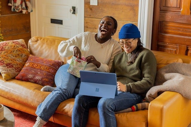 two people sitting on a couch laughing and looking down at a tablet