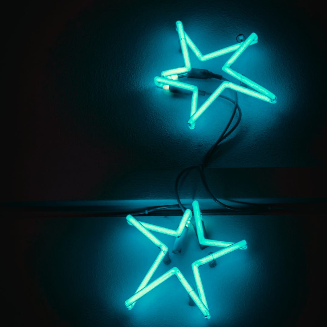 Against a dark background, two bright blue neon lights shaped like stars glow, lighting up the area in around them Against a dark background, two bright blue neon lights shaped like stars glow, lighting up the area in around them