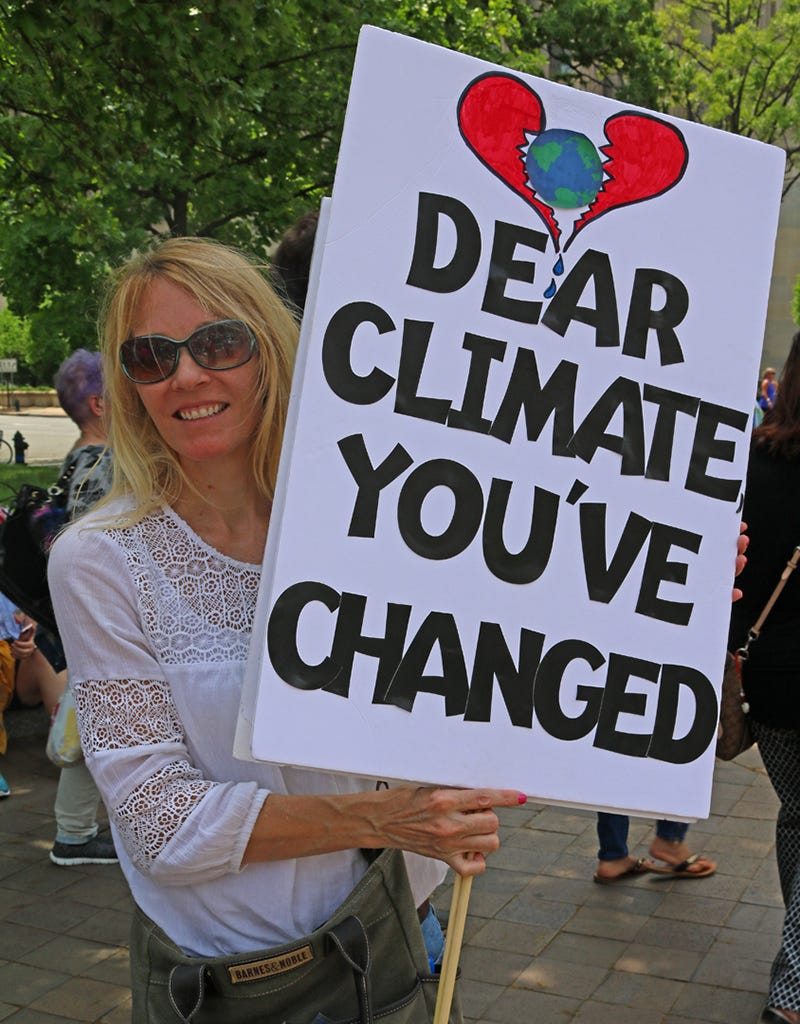 Photo of a woman at a protest march, holding a sign that says "Dear Climate, You've Changed"