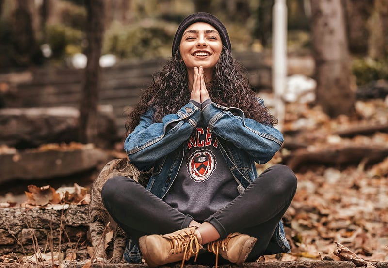 A smiling woman, in a meditative pose, sitting in the woods.