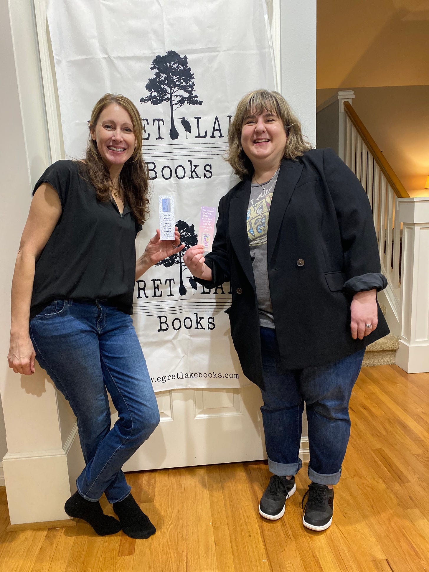 Celaine (left) and Amy (right) stand in front of the Egret Lake Books banner, holding up bookmarks for each of their books. They are both wearing jeans and black tops.