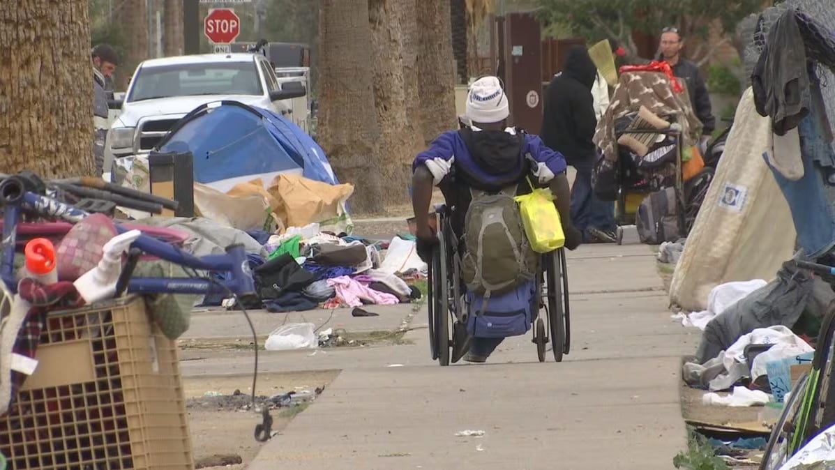 Homeless person in wheelchair rolls away from the camera down sidewalk, tents and shopping carts holding their meager possessions.