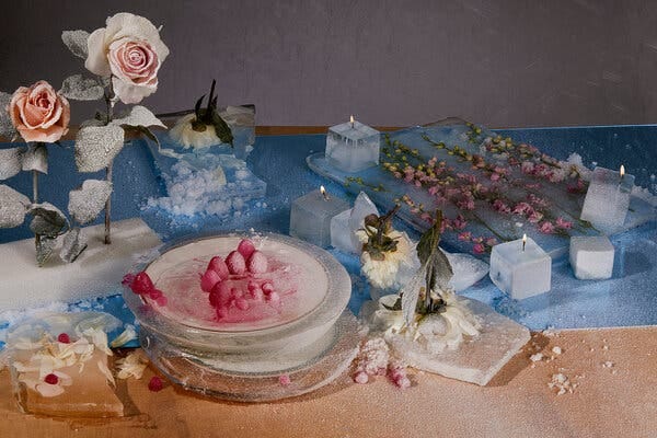 Two flowers covered in rime, balanced upright with their stems in foam; a few lit candles resembling ice cubes; a long sheet of ice with small flowers encased inside; and a bowl filled with ice, with strawberries and raspberries made of ice on top, stacked on a frosted plate.