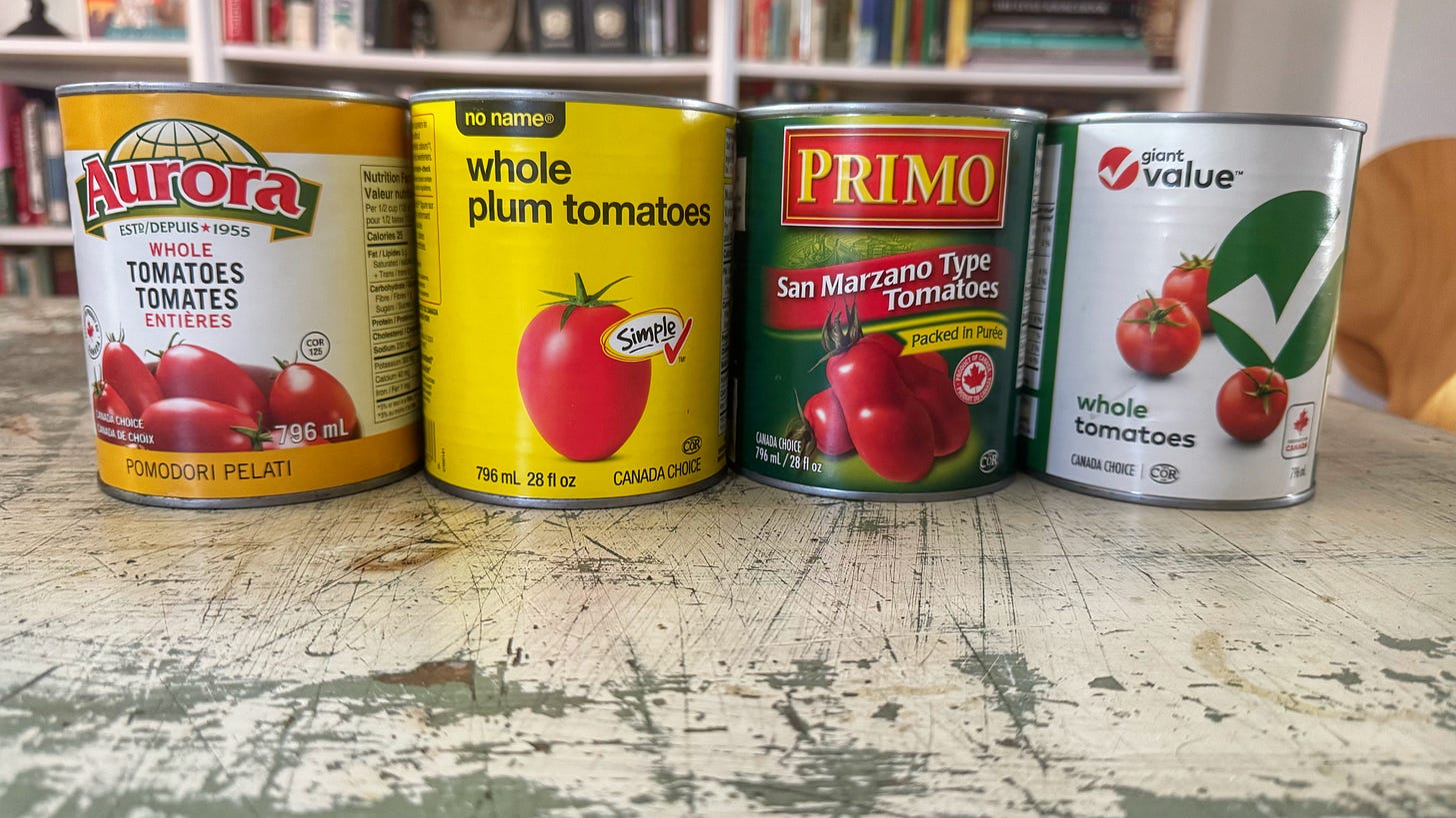 Cans of tomatoes on a dining room table. From left to right: Aurora Whole Tomatoes, No Name Whole Plum Tomatoes, Primo San Marzano Style Tomatoes, Giant Value Whole Tomatoes
