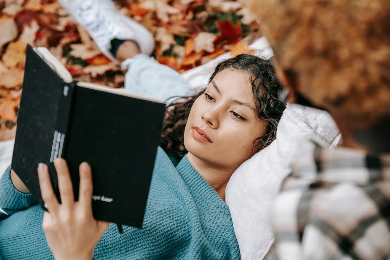 A woman lays on a blanket in a bed of autumn leaves, reading a book she holds in her hands.