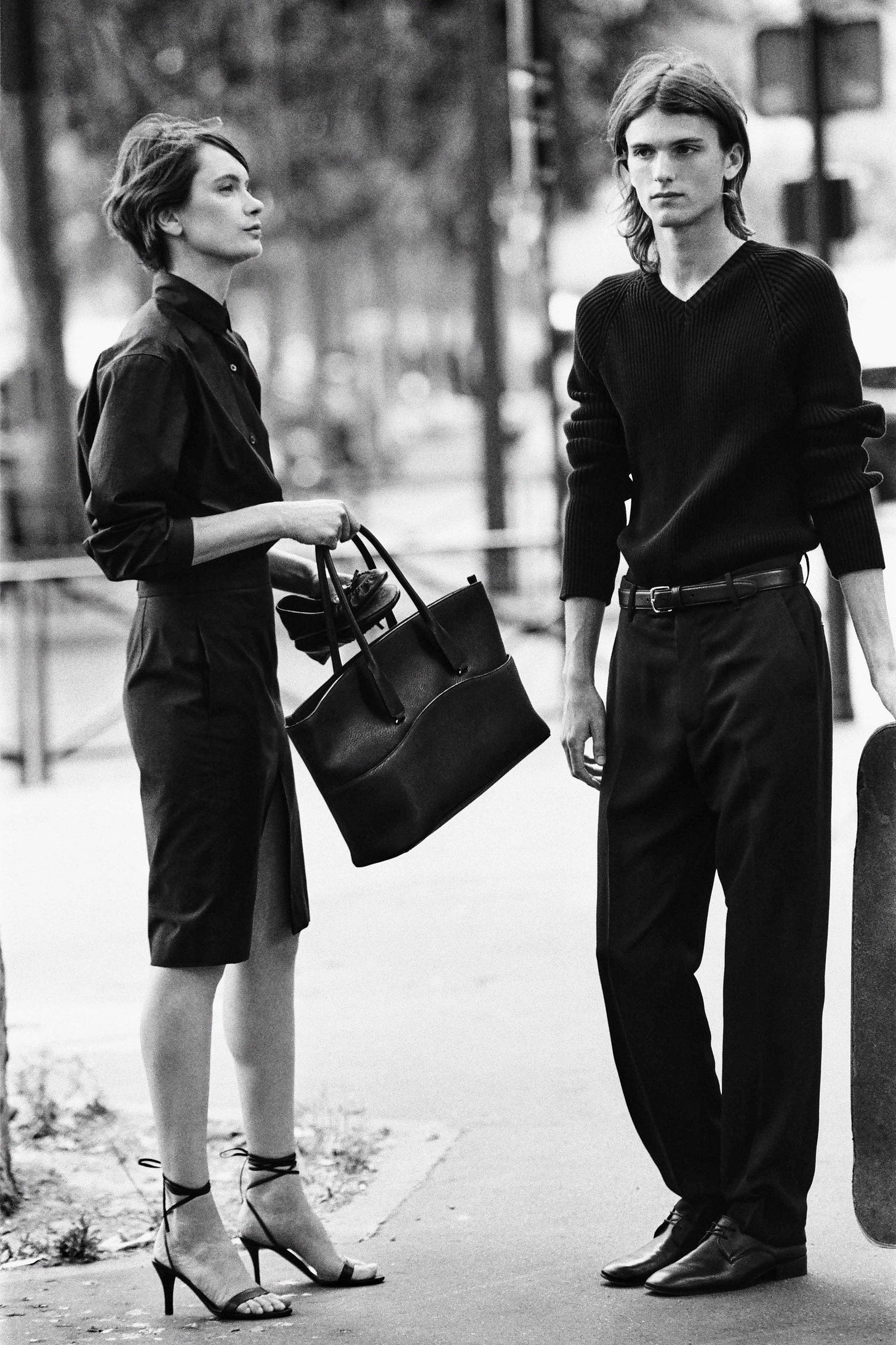 A black and white image depicts a woman and a man standing next to one another, both in completely black garb. The woman wears a tailored button-up shirt and a tilored pencil skirt while holding a leather tote and wearing strappy heels. The man wears a slim ribbed sweater and wide-leg trousers held with a slim belt and derby shoes.