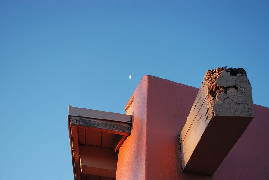 The corner of an adobe style picnic pavilion is highlighted by early morning light with the moon still showing in the sky above.