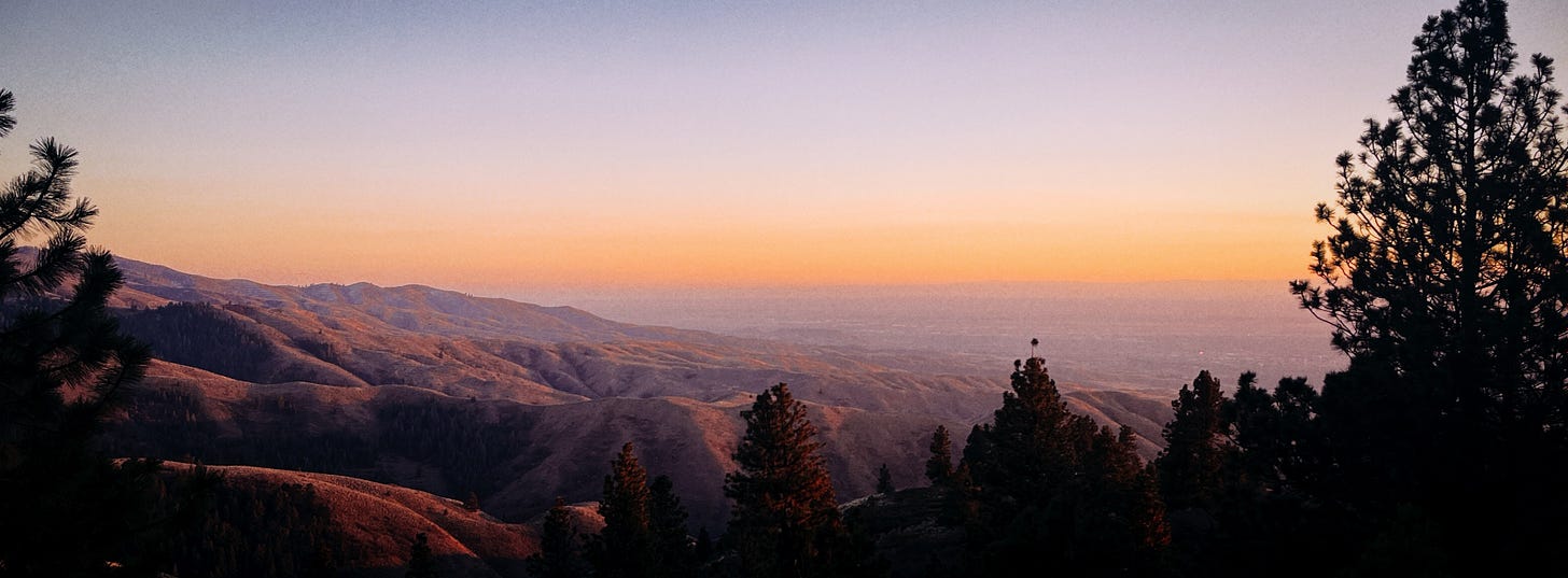 A wide-angle photograph taken from a mountain above Boise, Idaho. Hills stretch out in the distance. They look purple because of the sunset, which glows orange on the horizon.