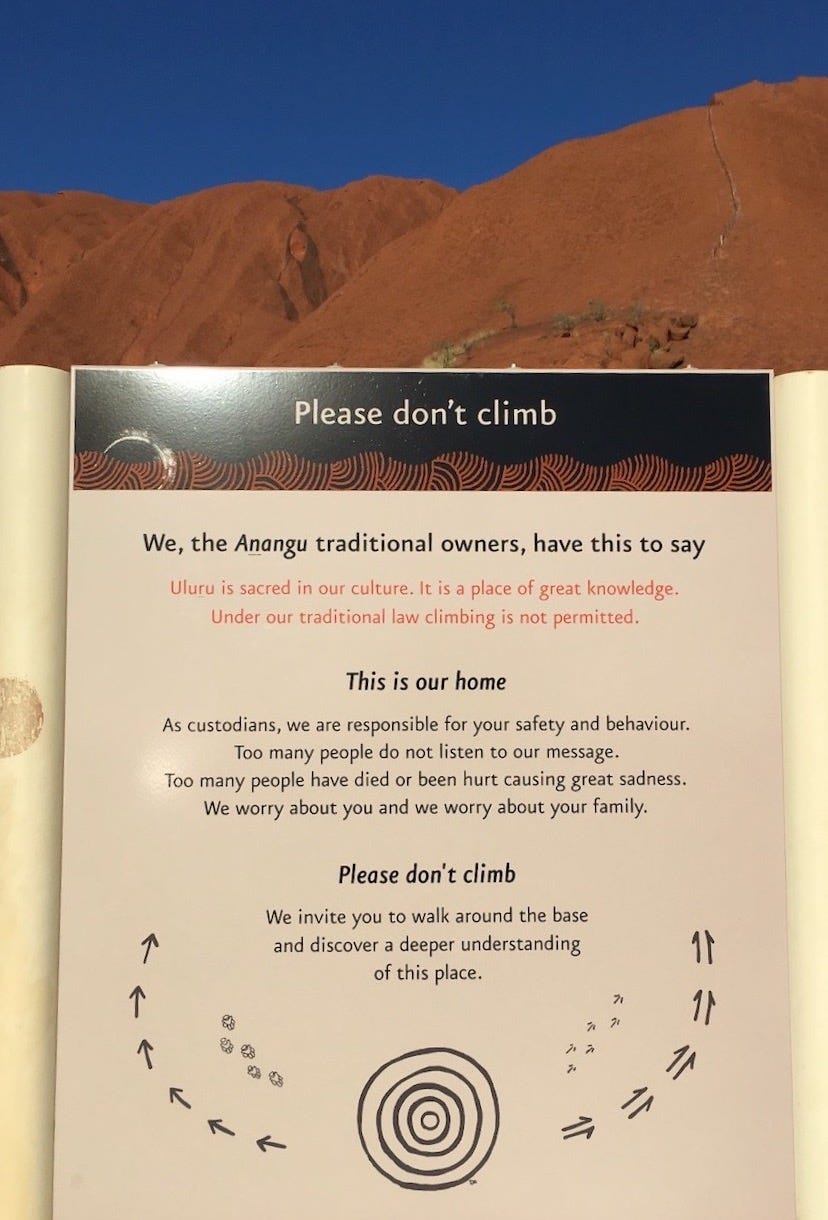 Sign in front of mountain and blue sky with text: “We, the Anangu traditional owners have this to say: Uluru is sacred in our culture. It is a place of great knowledge. Under our traditional law climbing is not permitted. This is our home. As custodians, we are responsible for your safety and behaviour. Too many people do not listen to our message. Too many people have died or been hurt causing great sadness. We worry about you and we worry about your family. Please don’t climb. We invite you to walk around the base and discover a deeper meaning of this place.” 