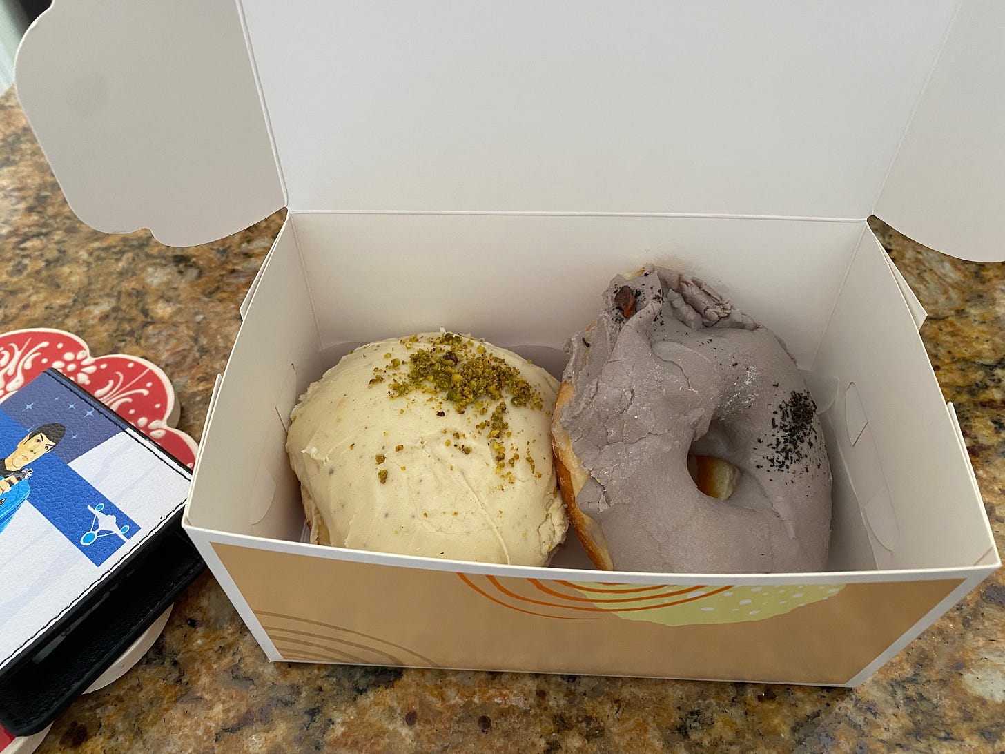 Two donuts in a white box with tan, orange, and yellow designs on the outside. The left donut is a filled one, with white icing and crumbled pistachio. The right is a classic donut shape with light purple icing and crumbles of earl grey tea leaves. On the left side of the frame my Star Trek wallet is visible.