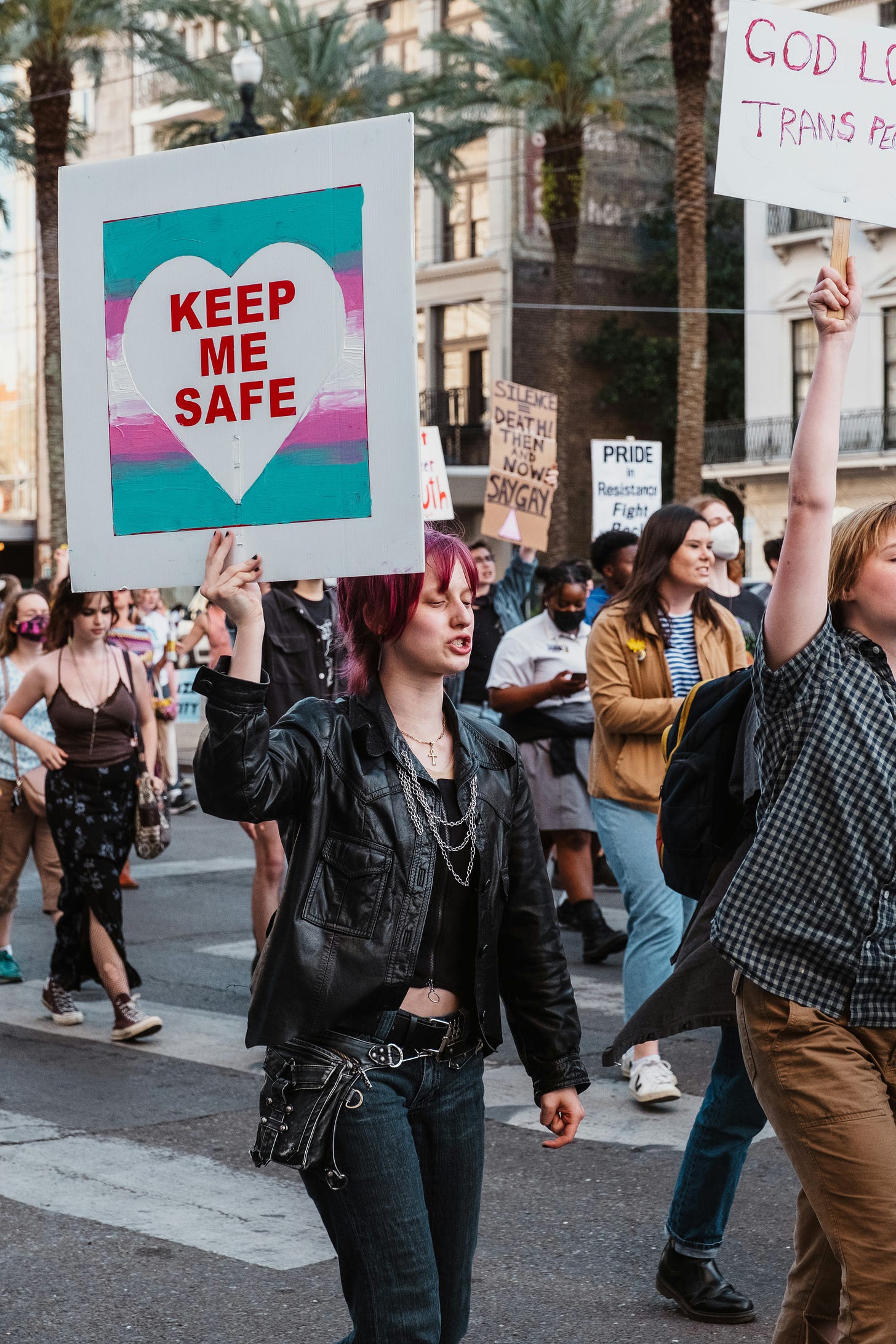 A person with red hair holding up a sign that reads "KEEP ME SAFE" with a heart symbol during a protest march, with other demonstrators and signs in the background.