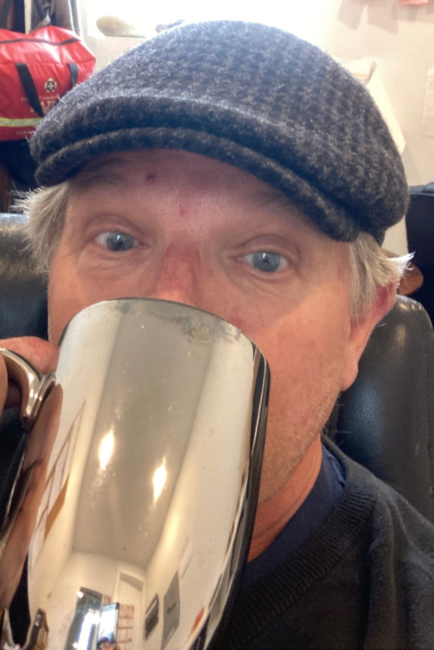 The author Bradley James Cooper, has a chrome coffee mug at his mouth, above the rim are his eyes in an expression of mild surprise and he's wearing a tweed cap.