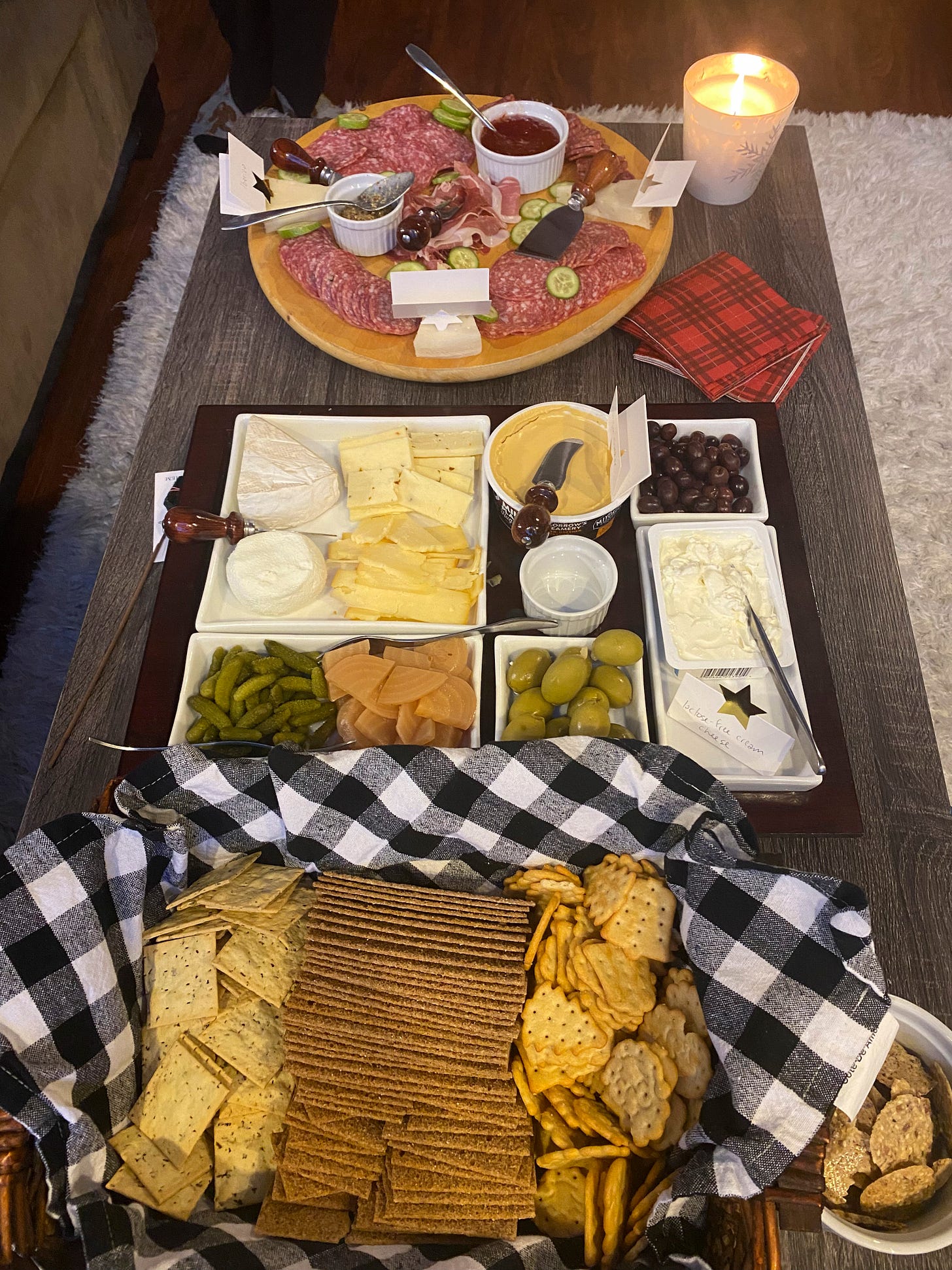 A coffee table with charcuterie. Furthest away is a wood lazy susan with meats, cucumber slices, Spanish cheeses, and ramekins of mustard and jam. In the middle is a divided serving platter with several types of cheese, olives, and pickles. In the foreground is a basked of three different types of crackers, lined with a black-and-white gingham napkin.