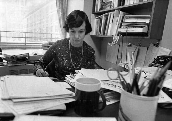 A black-and-white photo of Betty Prashker, a woman wearing a dress and pearls, sitting at a cluttered desk with a pencil in her right hand.