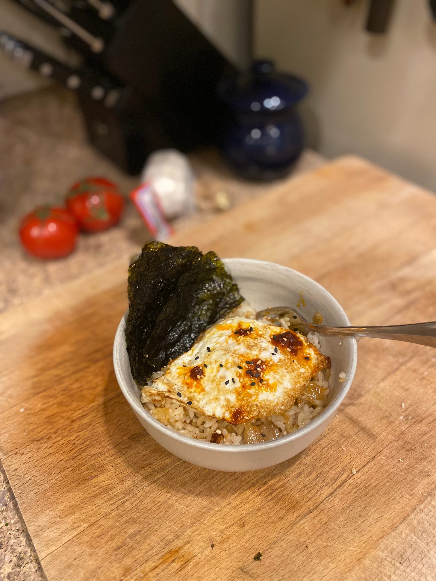 Sitting on a wood cutting board, an off-white bowl of fried rice topped with an over easy egg. It's garnished with chili miso and two colours of sesame seeds, and a few sheets of gim fanned at the side of the bowl. A fork sticks out on the right hand side.