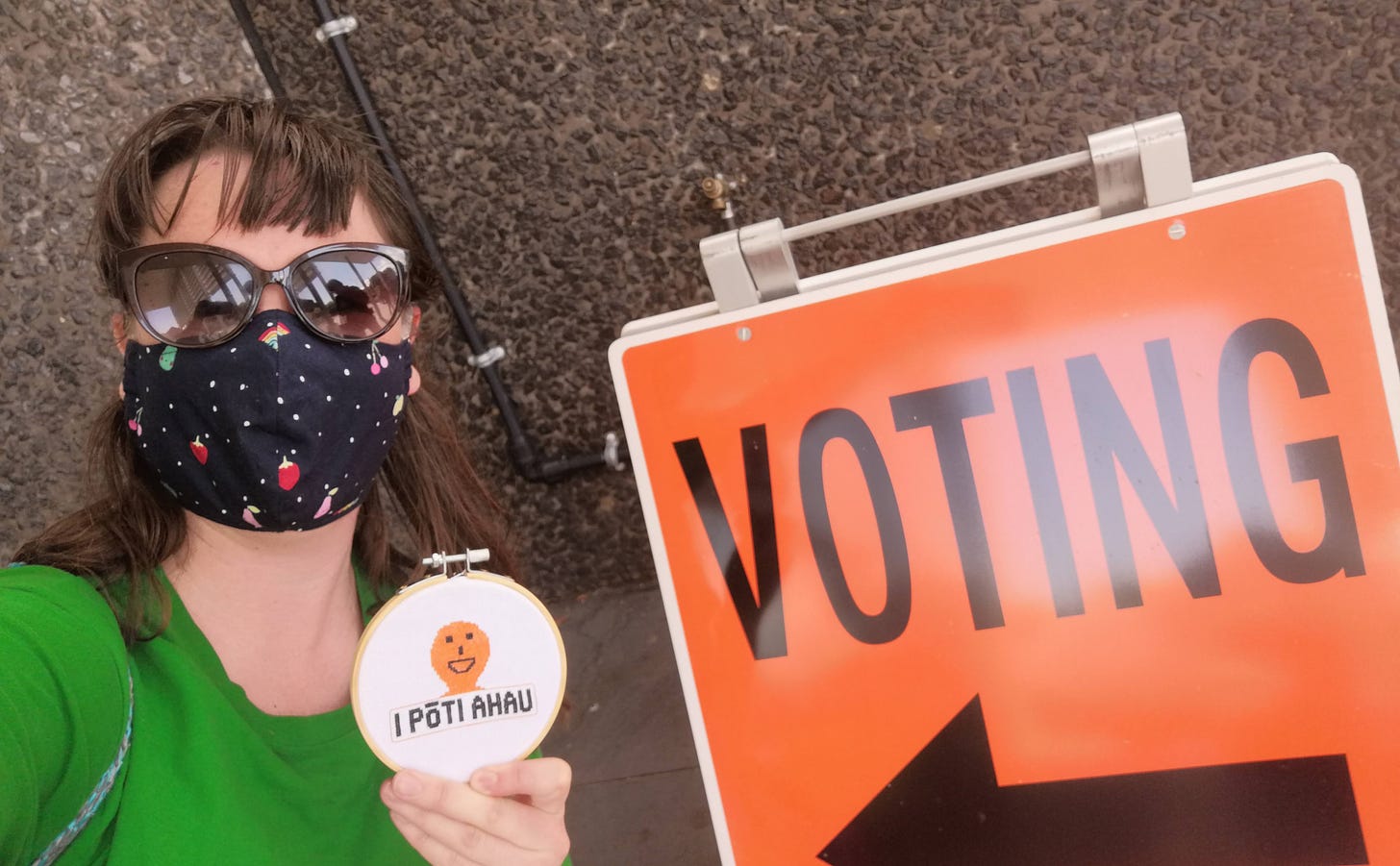 A white woman wearing a fabric face mask and sunglasses beside an orange "VOTING" sign holding a cross stitch in a frame with the orange election guy and the text " I PŌTI AHAU"