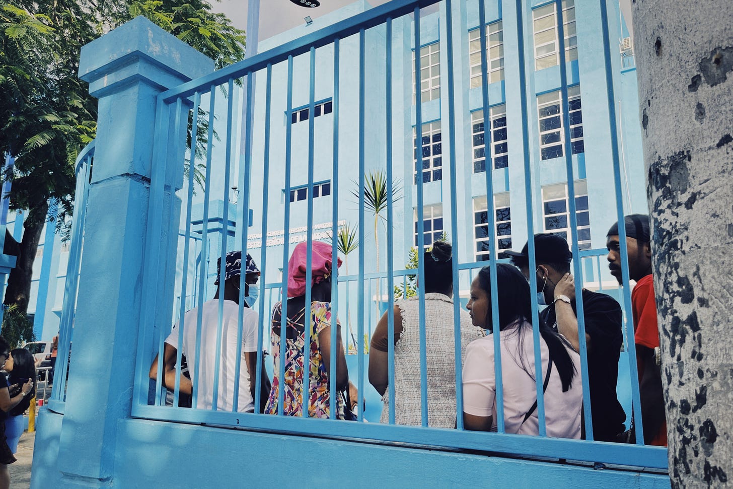 Black people wait in line in a fenced-in area outside a hospital in Santo Domingo. Both the hospital, visible in the background, and the fence, in the foreground, are painted bright blue.