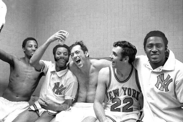 A black-and-white photo of five men, three of them wearing Knicks jerseys and two of them shirtless, sitting in a locker-room shower, their arms around one another, with giddy expressions on their faces.