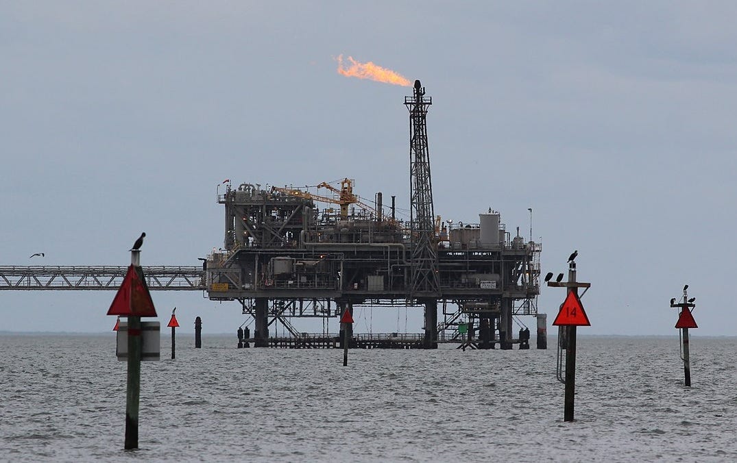 Against a grey sky, in calm seas, an offshore oil platform flares methane from oil drilling. In the foreground, cormorants perch on marker signs 