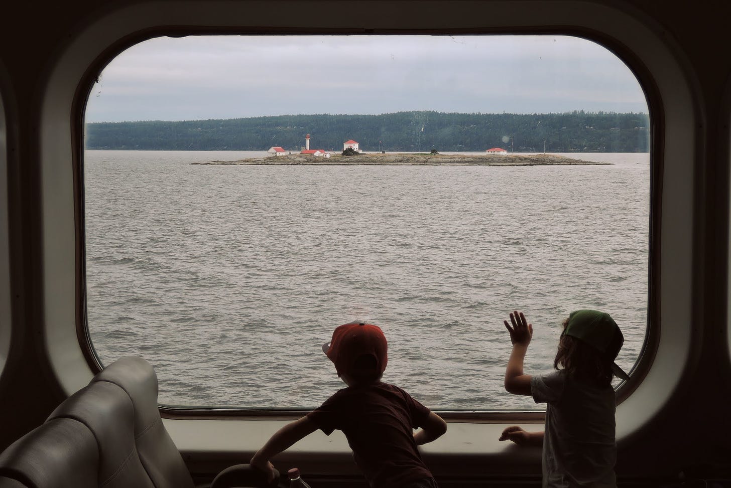 Two children look out the window of the Vancouver-Nanaimo ferry towards a small island with a lighthouse.