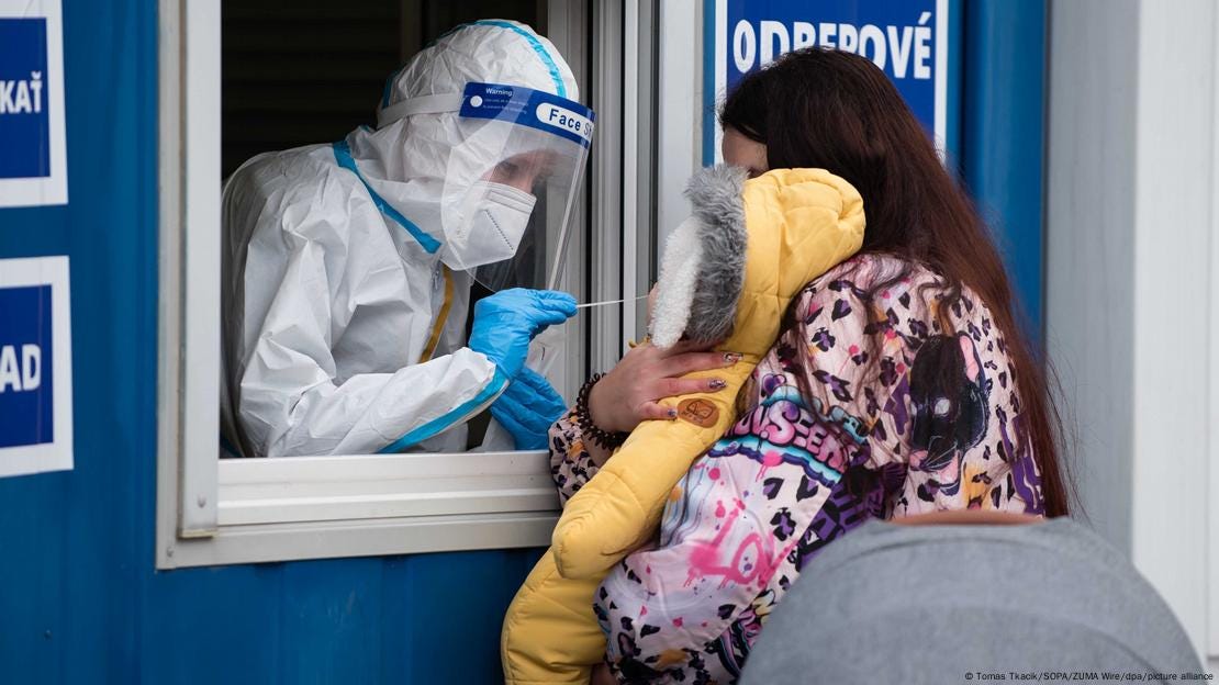 A healthcare worker in protective clothing takes a nasal swab sample from a toddler during mass corona testing. The child is held by his mother to the window behind which the healthcare worker is standing.