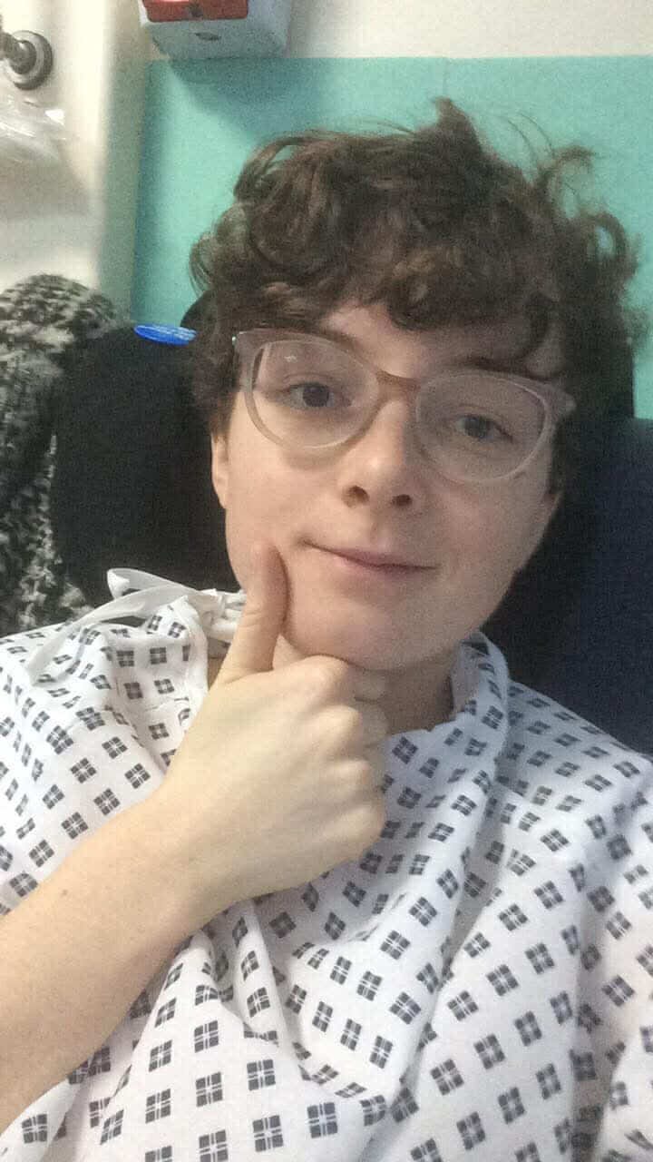 A picture of Rachel, a white person with short curly hair and light pink glasses, wearing a surgery gown and putting her thumbs up