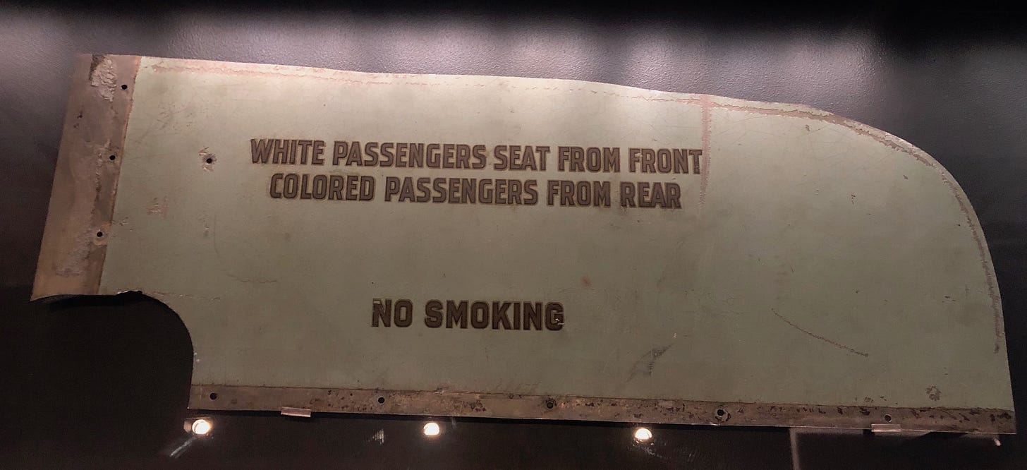A flat piece of metal bearing the words, “WHITE PASSENGERS SEAT FROM FRONT / COLORED PASSENGERS FROM REAR / NO SMOKING”.