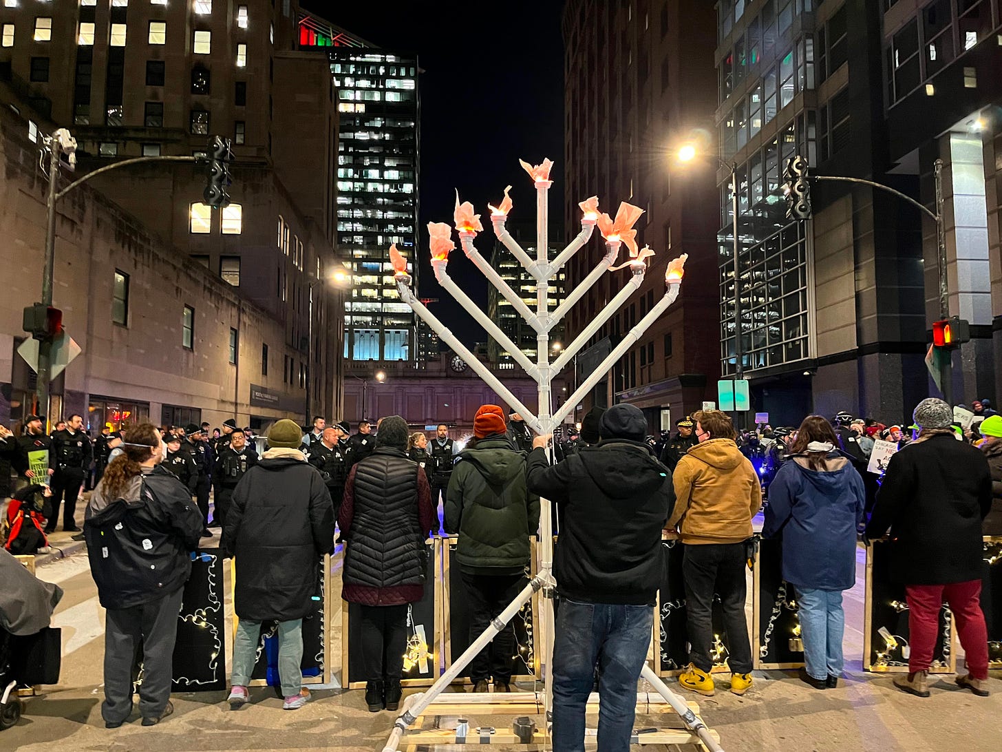 A photo of protesters holding lighted letters while blocking a street, taken from behind. There is a large menorah behind them.
