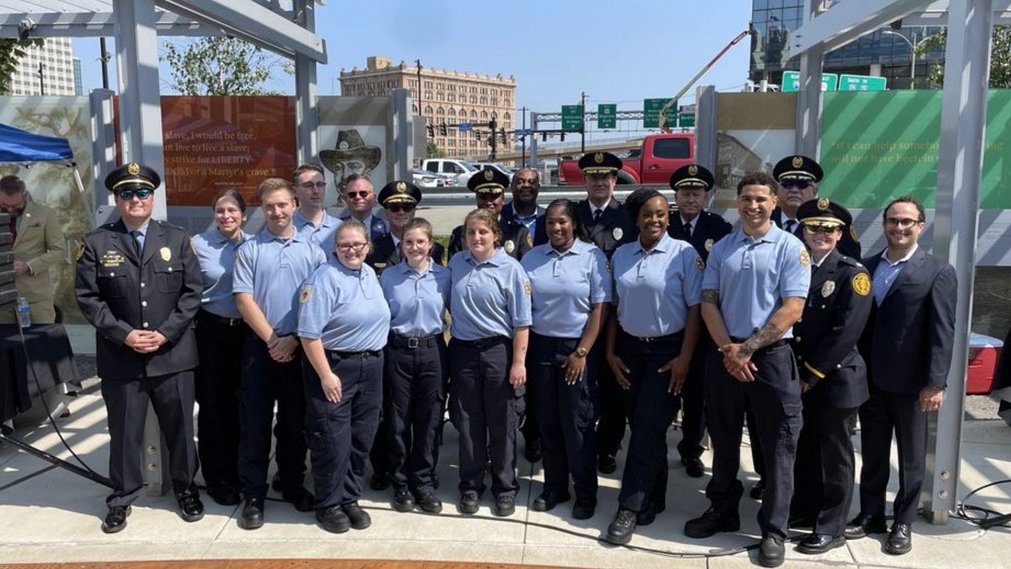 A group shot of nine blue-shirted paramedic graduates flanked by various officials in emergency response uniforms on a sunny day 