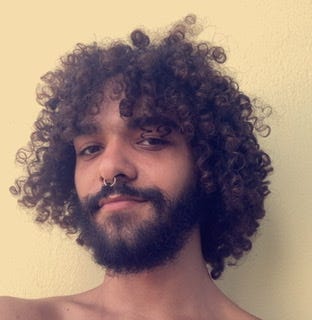 Warm skinned person with loose curls, a septum ring, and a beard in front of a yellow backdrop.